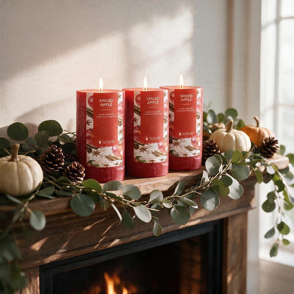 Three lit red Spiced Apple pillar candles on a fireplace mantel with eucalyptus garland, pinecones and white pumpkins