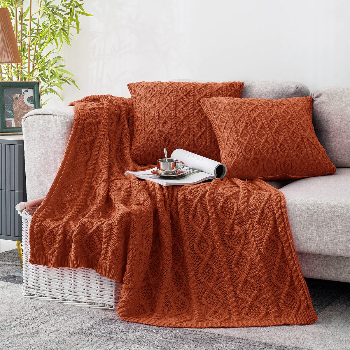 Rust cable knit throw blanket and two matching pillow covers on a light grey sofa with a magazine and teacup