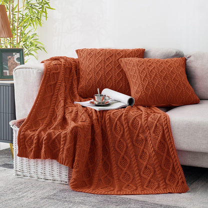 Rust cable knit throw blanket and two matching pillow covers on a light grey sofa with a magazine and teacup