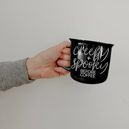 Hand holding the black Creepy and Spooky Before Coffee mug against a white wall