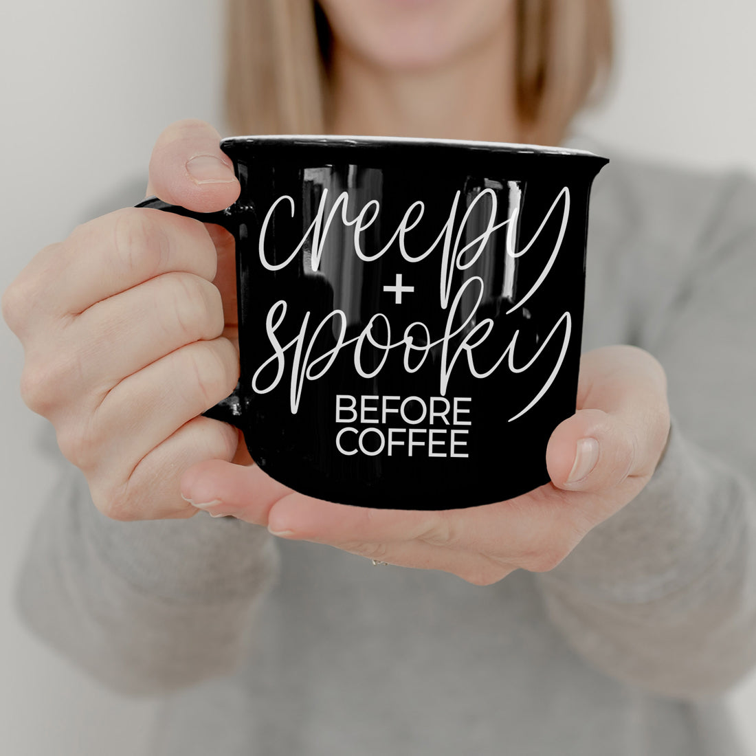 Hands holding the black Creepy and Spooky Before Coffee camp mug
