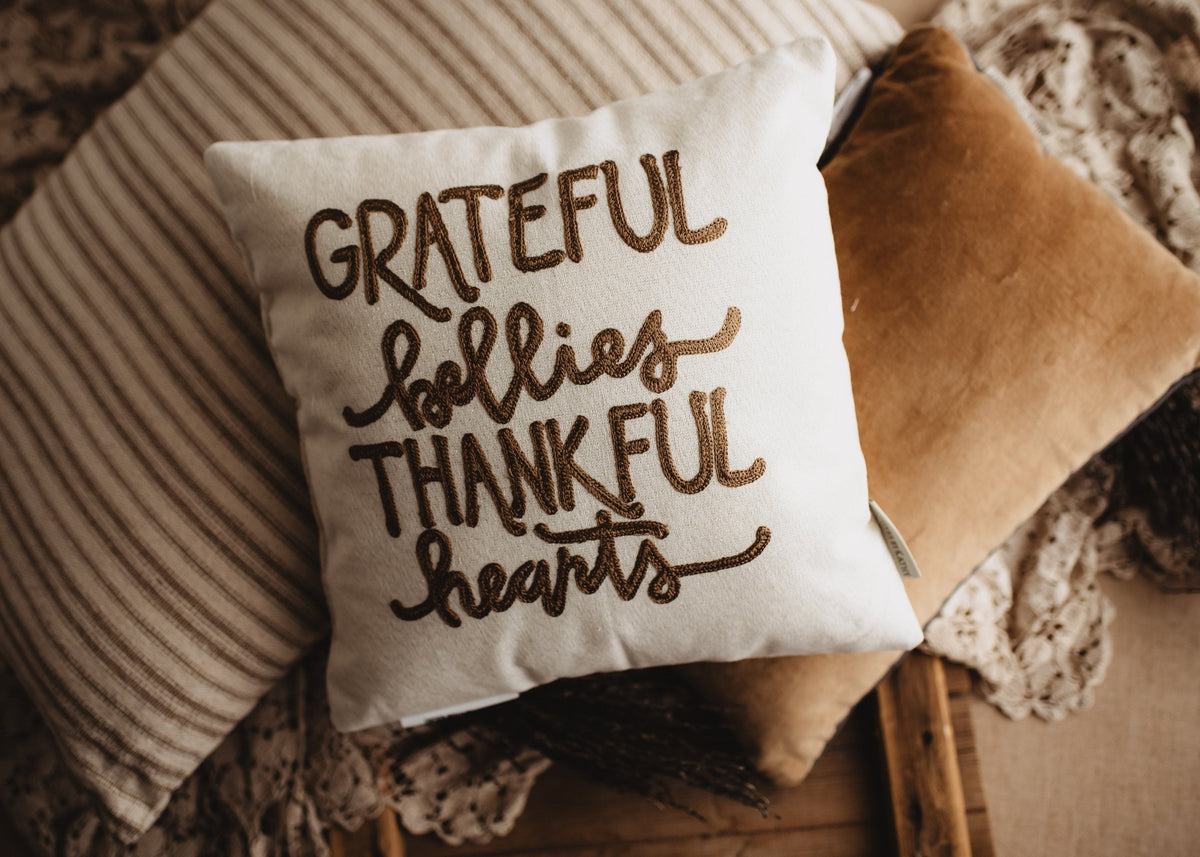 Grateful Bellies Thankful Hearts pillow on a bench with a striped blanket and tan pillow