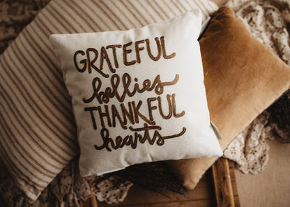 Grateful Bellies Thankful Hearts pillow on a bench with a striped blanket and tan pillow
