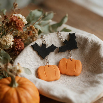 Bat and pumpkin drop earrings on a linen dish beside dried flowers and a mini pumpkin