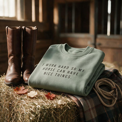 Sage green I Work Hard So My Horse Can Have Nice Things sweatshirt folded on a hay bale in a barn with riding boots and a rope halter