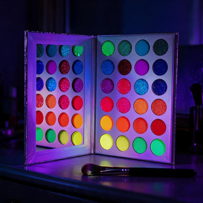 24-color neon eyeshadow palette with mirror glowing under blacklight beside a makeup brush