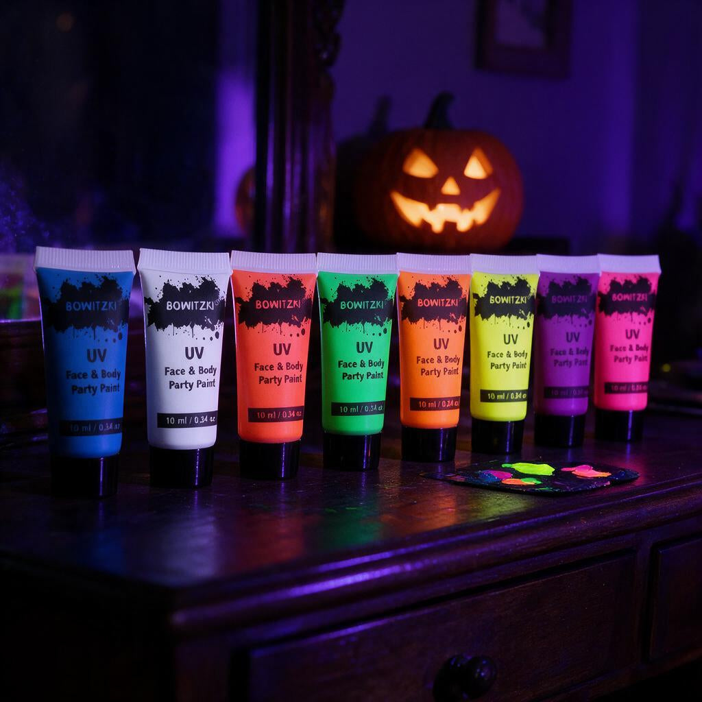 Eight UV neon face and body paint tubes glowing under blacklight on a dark vanity with a jack-o-lantern