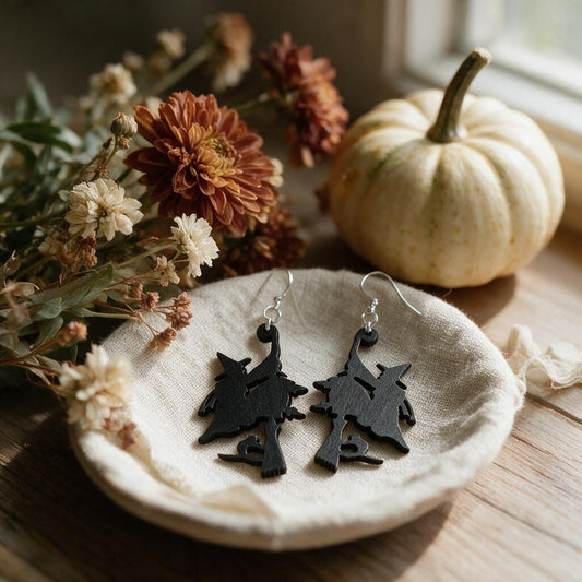 Black wooden witch on broomstick earrings on a linen dish beside a white pumpkin